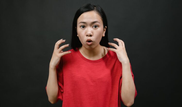 Asian woman in a red shirt expressing surprise with hands raised, on a dark background.