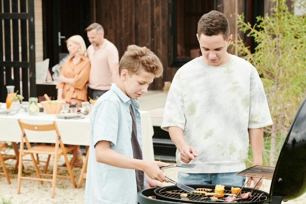 Two boys grilling at a family barbecue with parents in the background.