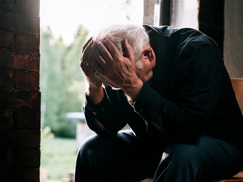 Senior man in black, grieving alone, sitting on a wooden chair by a doorway in a pensive pose.