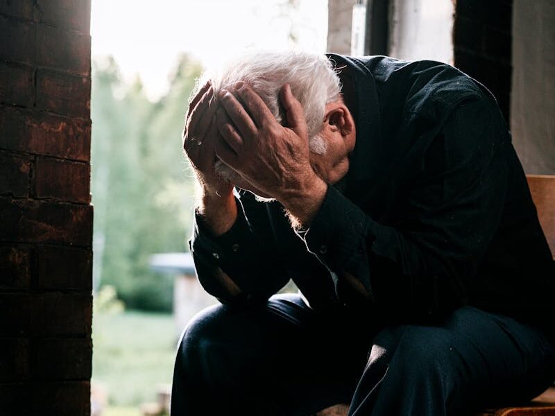 Senior man in black, grieving alone, sitting on a wooden chair by a doorway in a pensive pose.