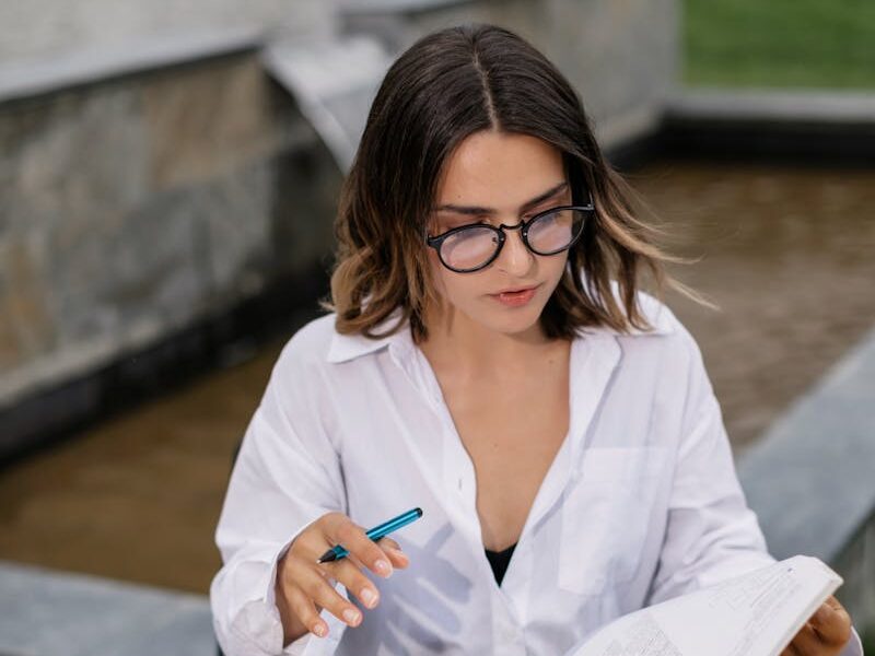 A young woman with eyeglasses studying outdoors with books and a laptop, portraying a studious and focused atmosphere.