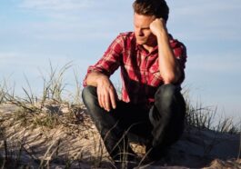Man in a red plaid shirt sitting thoughtfully on a sandy dune in Australia, surrounded by grass.