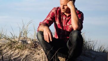 Man in a red plaid shirt sitting thoughtfully on a sandy dune in Australia, surrounded by grass.