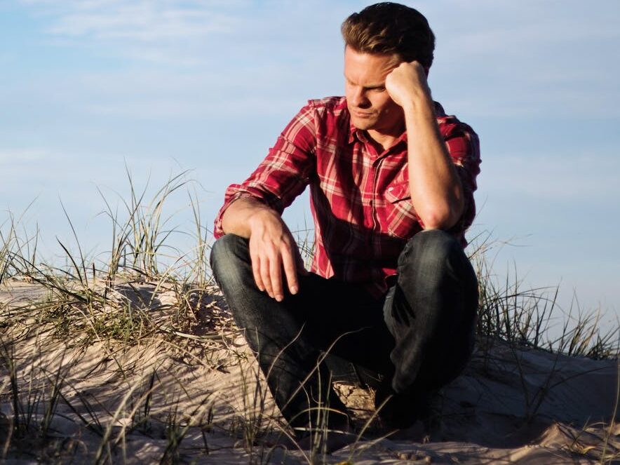 Man in a red plaid shirt sitting thoughtfully on a sandy dune in Australia, surrounded by grass.