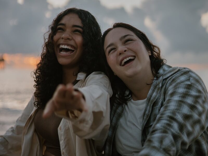two women sitting on the beach pointing at the camera