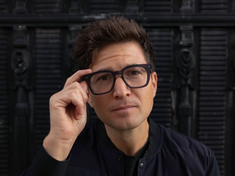 a man wearing glasses sitting in front of a black wall