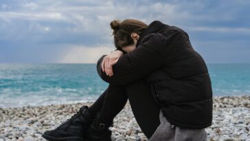 a woman sitting on a rocky beach with her head in her hands