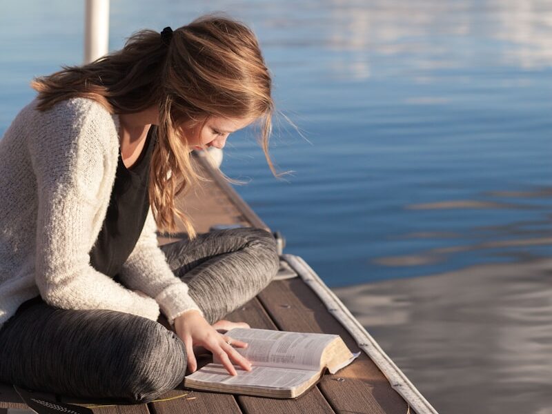 photo of woman reading book near body of water