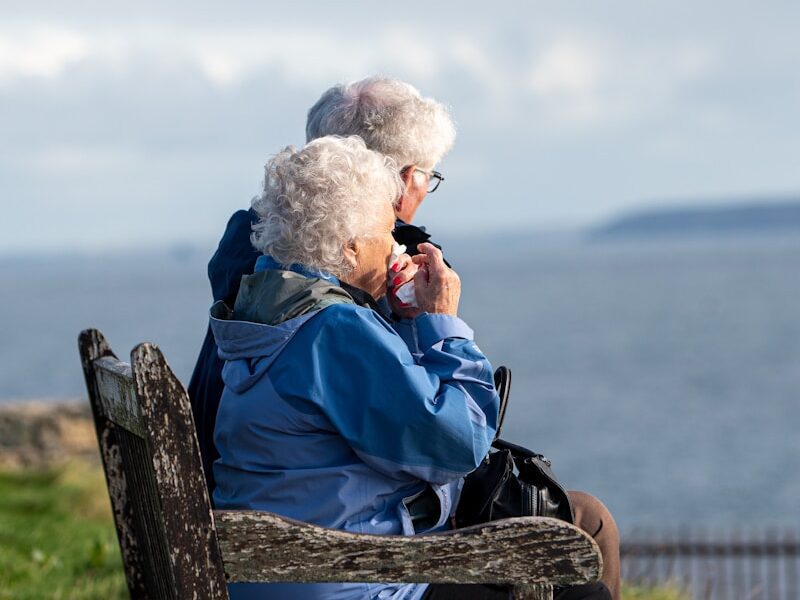man and woman sitting on gray wooden bench viewing blue sea during daytime