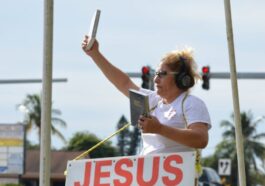 A woman stands on the roadside holding a Bible and a sign promoting Christian faith.