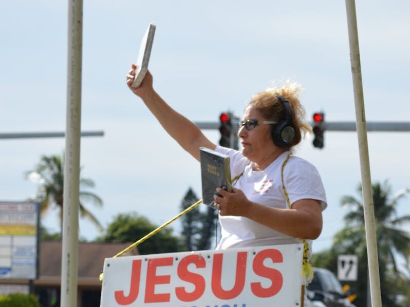 A woman stands on the roadside holding a Bible and a sign promoting Christian faith.