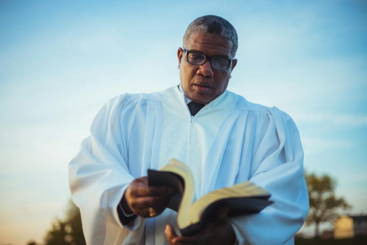 An adult pastor wearing glasses and a white robe reads a book outdoors under a blue sky.