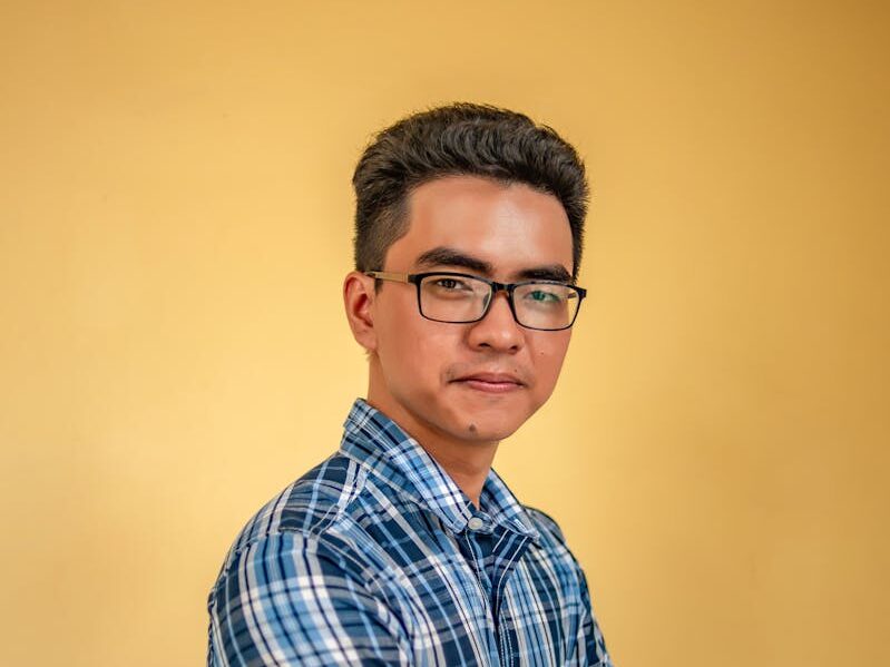 A portrait of a confident young man in glasses wearing a plaid shirt against a neutral background.