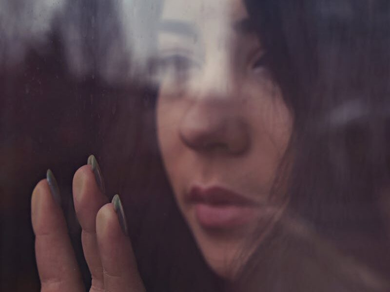 A thoughtful young woman looks out a window, her reflection showing emotion.