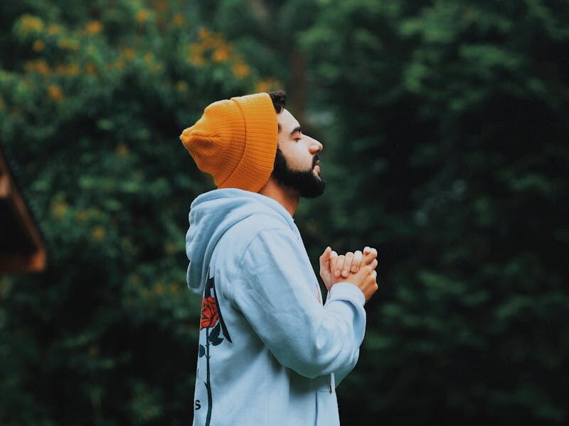 A man in a beanie and hoodie deeply praying outside in a lush green forest.