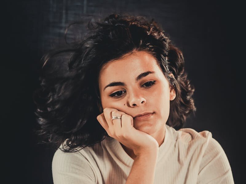 Portrait of a woman sitting pensively, wearing a casual white top. Moody, introspective atmosphere.