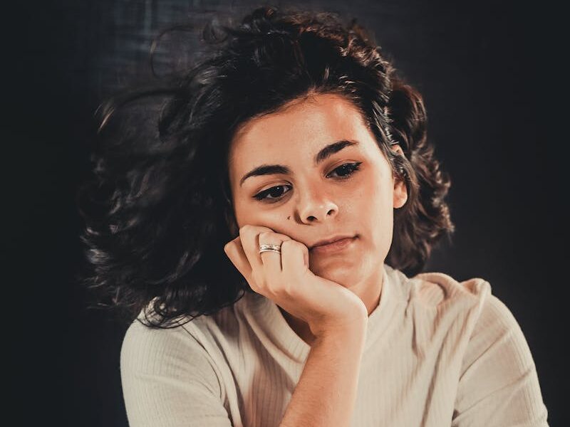 Portrait of a woman sitting pensively, wearing a casual white top. Moody, introspective atmosphere.