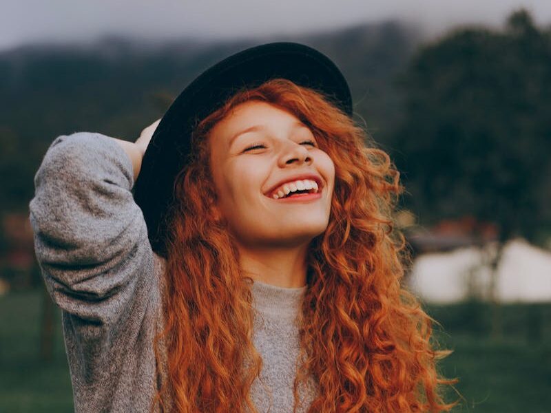 A happy young woman with curly red hair and a hat smiling in a lush green park.