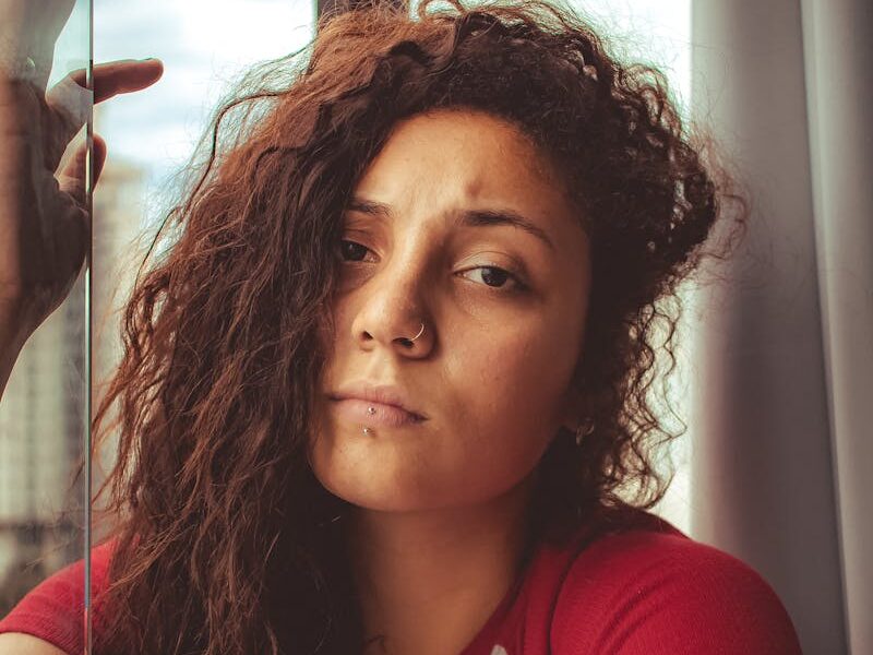 A woman with curly hair looking thoughtfully outside a window in a red striped shirt.