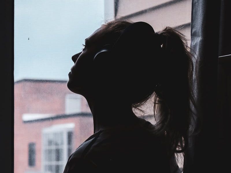 A silhouette of a woman wearing headphones, deep in thought by a window.