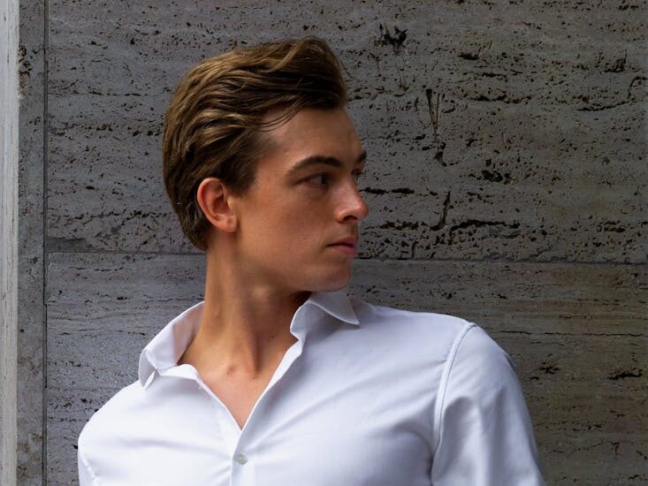 Confident young man in white outfit posing against a concrete wall.