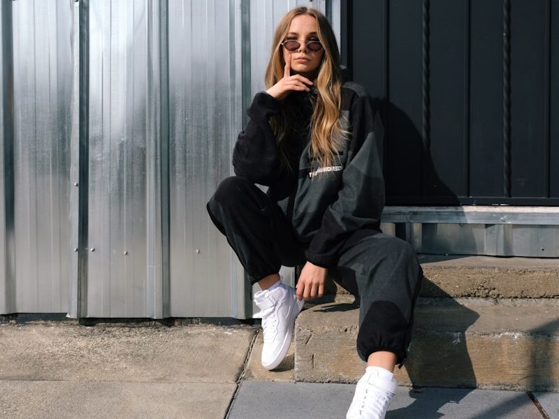 woman in black jacket and white pants sitting on gray concrete floor