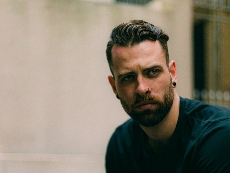 Portrait of a stylish, tattooed man leaning on a railing with a contemplative expression.