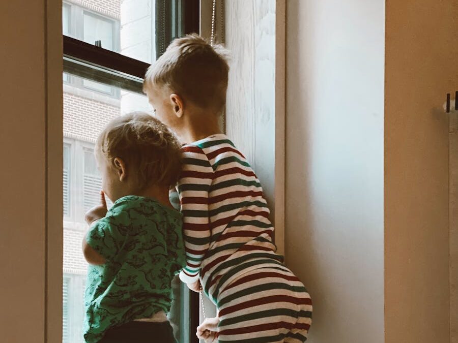 Two kids in pajamas curiously peering out of a window inside their home.