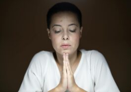 A peaceful woman with closed eyes and clasped hands in deep prayer inside a quiet room.