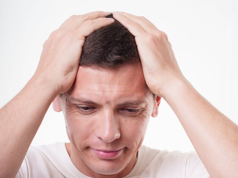 A young adult man in a white shirt appears stressed, holding his head with both hands against a plain background.