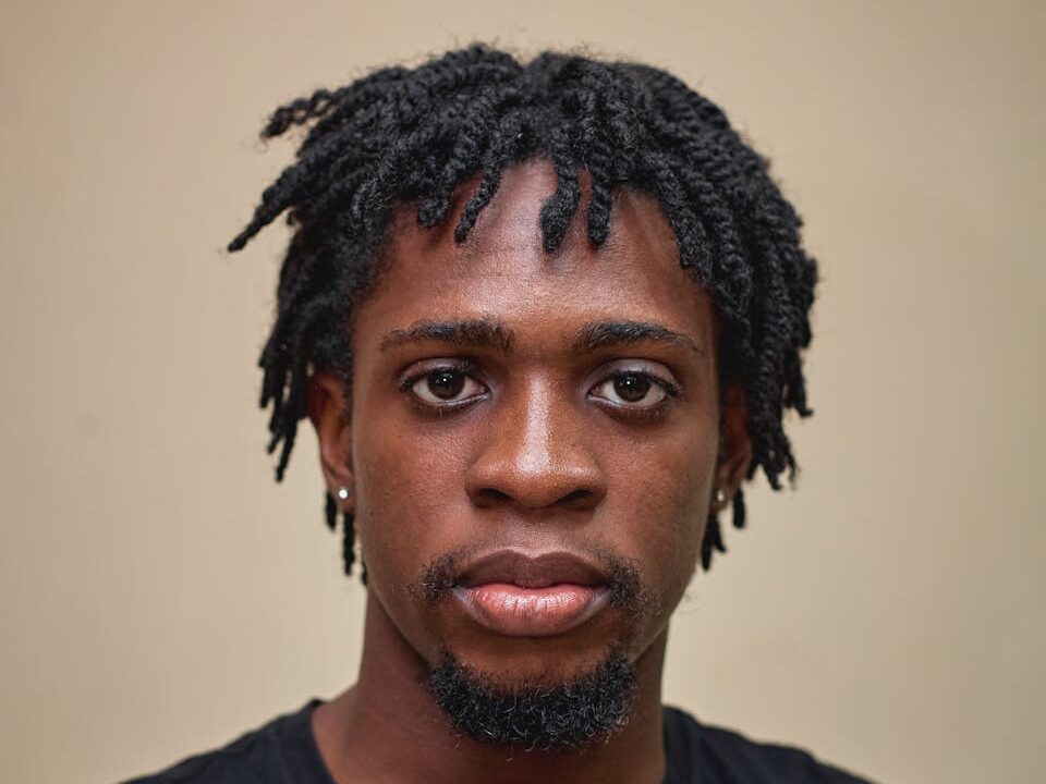 Close-up portrait of a young man with dreadlocks looking directly at the camera against a neutral background.