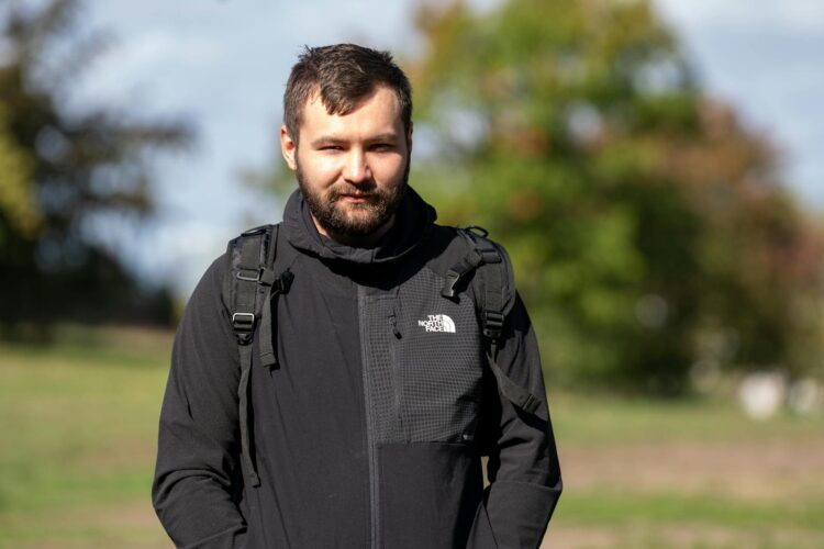 A man in a black jacket stands outdoors in a park during the day.