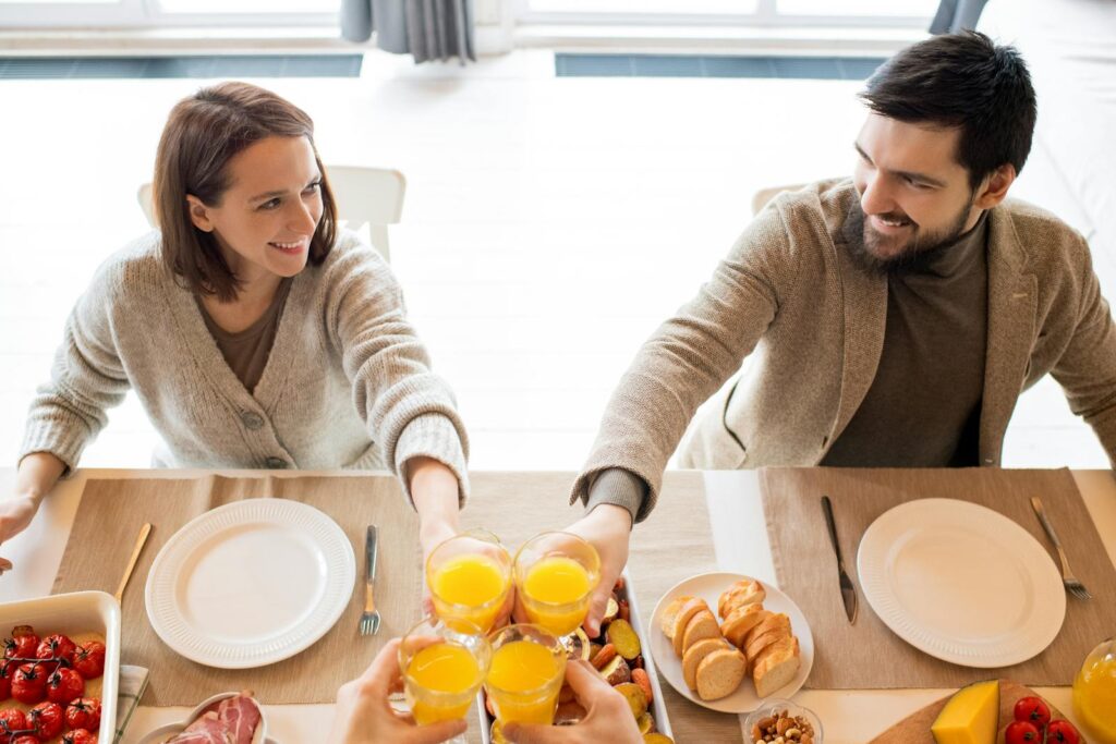 A joyful couple shares a toast with orange juice at a sunny breakfast table indoors.