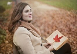 Side view of peaceful young female in warm coat and scarf sitting on bench with open book in hands and looking away pensively while resting in autumn park