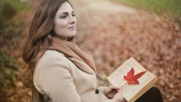 Side view of peaceful young female in warm coat and scarf sitting on bench with open book in hands and looking away pensively while resting in autumn park