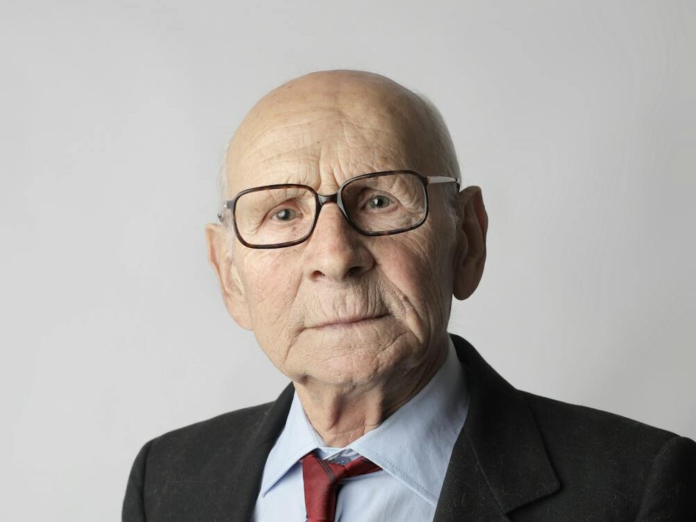 An elderly man with glasses holding dollar bills, representing wealth and financial security.