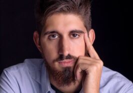 Close-up portrait of a young bearded man in a blue shirt, looking thoughtful.