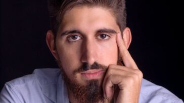 Close-up portrait of a young bearded man in a blue shirt, looking thoughtful.