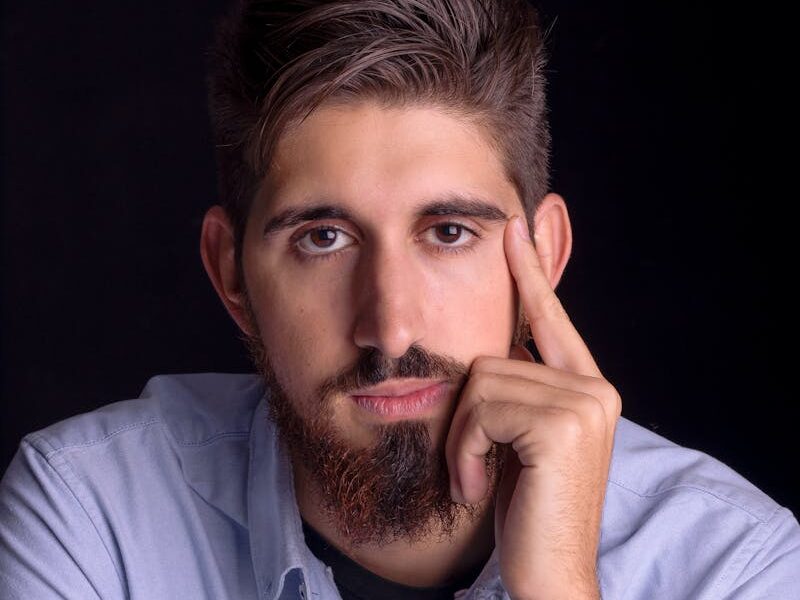 Close-up portrait of a young bearded man in a blue shirt, looking thoughtful.