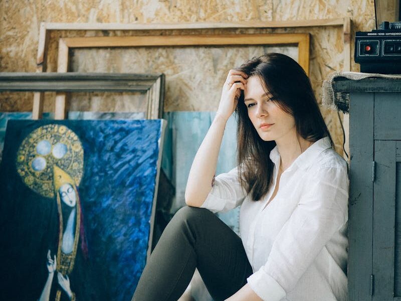 A woman sitting in an art studio, surrounded by paintings, deep in contemplation.