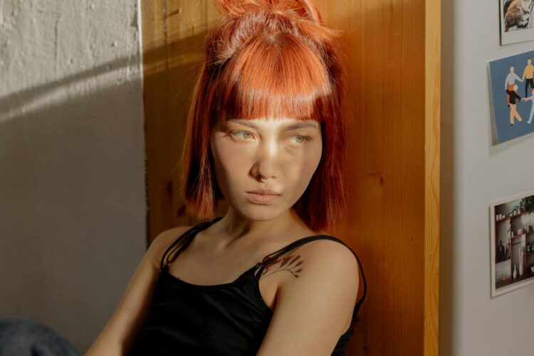 A thoughtful woman with red hair sits gazing in a sunlit room, lost in contemplation.