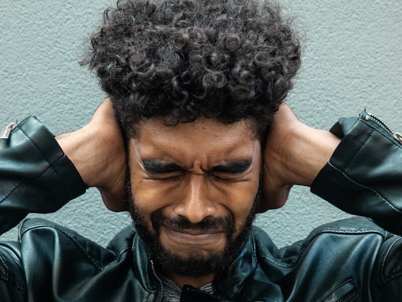 A man with curly hair and a beard shows frustration while covering his ears.