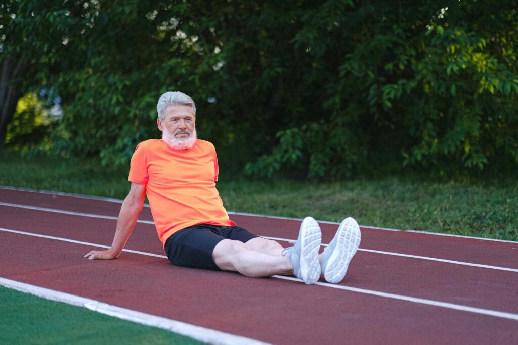 Full body of elderly satisfied male sitting and relaxing after tiring workout in stadium with lush shrubs and trees