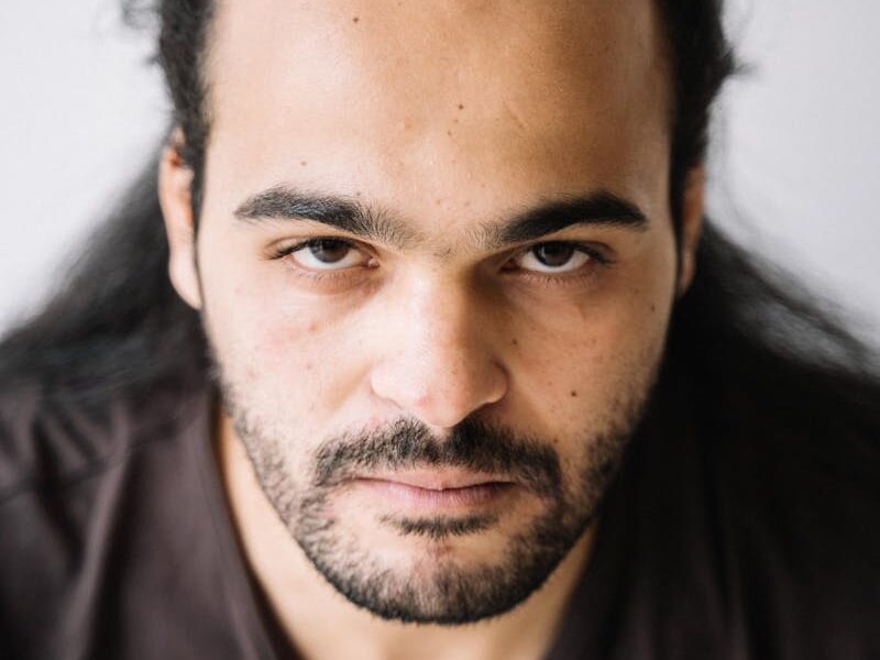 Close-up portrait of a bearded man with long hair wearing a crew neck t-shirt looking directly at the camera.
