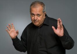 Studio portrait of an older man with a playful gesture in a black shirt.