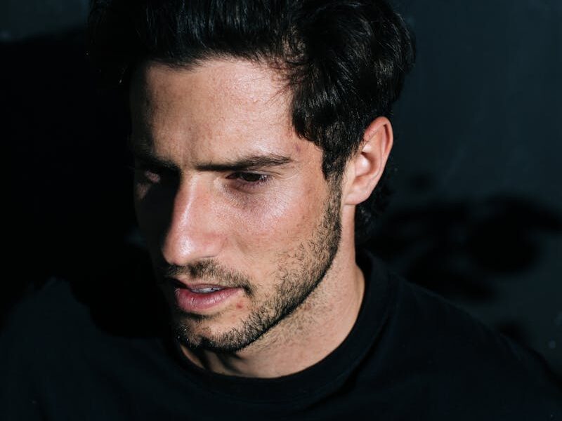 From above side view of pensive male with beard and dark hair looking away while standing on black background in bright sunshine