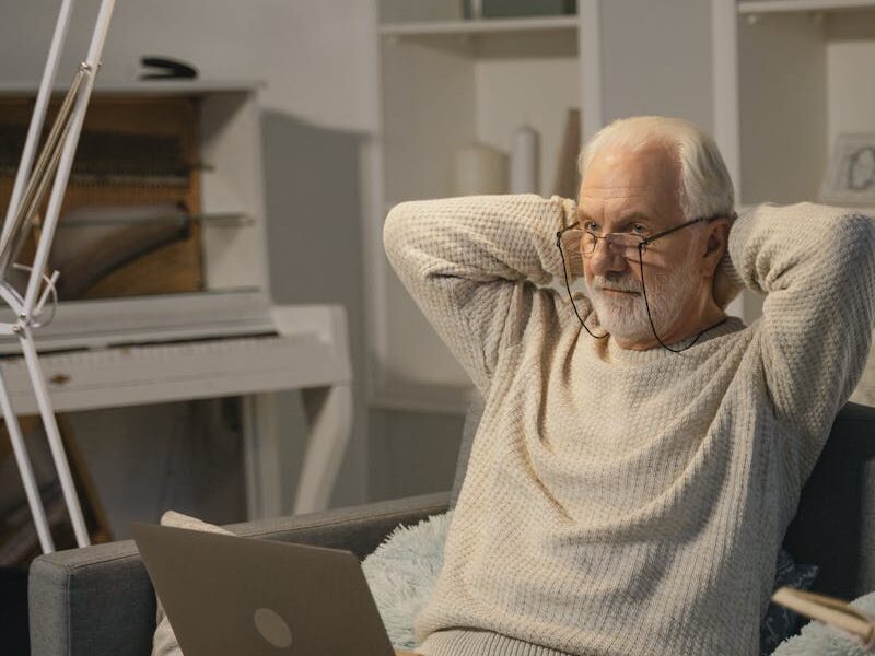 Elderly man sitting comfortably indoors using a laptop, wearing a knitted sweater.
