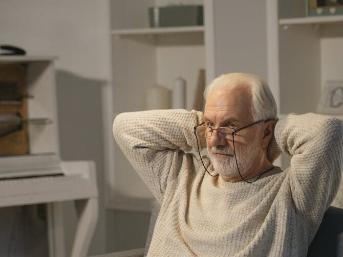 Elderly man sitting comfortably indoors using a laptop, wearing a knitted sweater.