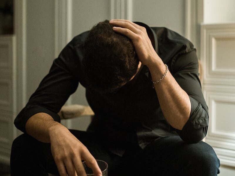 Man in distress seated indoors, head in hand, holding a drink, reflecting solitude.