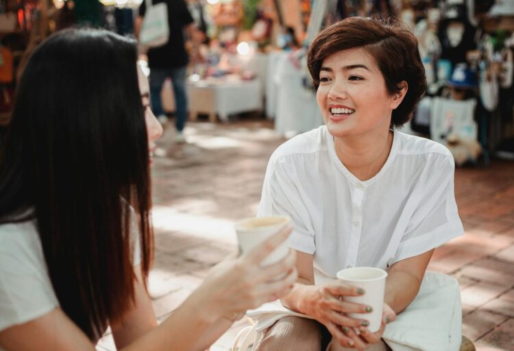 Two women engaged in cheerful conversation at an outdoor market, holding coffee cups.